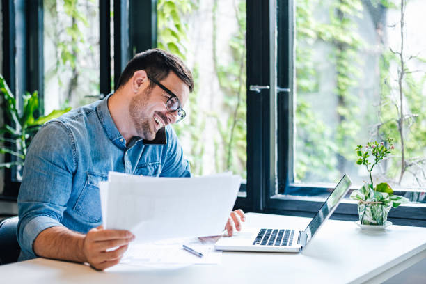 istock Businessman with documents discussing on smart phone while using laptop at home 1405602591