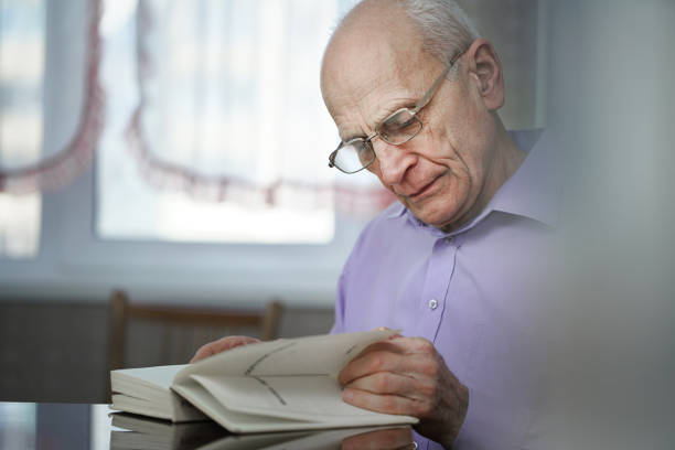 istock Lonely old man reads book seated at table at home 1405604337
