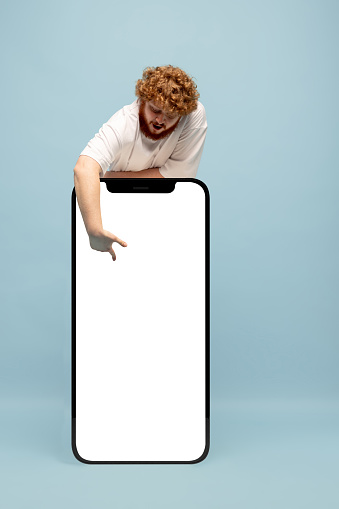 Pointing at device screen. Red-headed man standing next to huge 3d model of smartphone with empty white screen isolated on blue background, Recommending new app or website. Mockup for ad