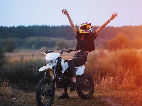 A beautiful young happy woman in a helmet poses sitting on a cross-country motorcycle at sunset