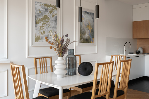 Stylish dining area with white table, six wooden chairs and decorations, modern lamps and interesting art next to simple kitchen