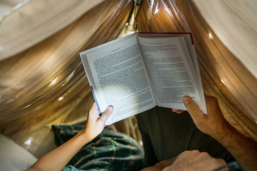 Portrait of a 6 year old boy and his father reading a book in teepee tent. Father and son with flashlight reading book under blanket at home.