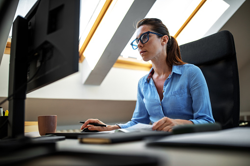 A beautiful young woman working at her home office on the desktop computer and focusing on a paper with numbers