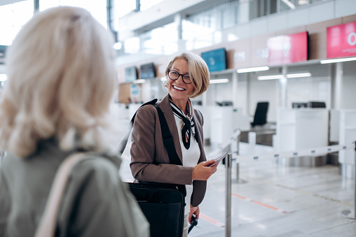 Two positive and charming women standing at modern airport terminal and waiting for passport check-up and scheduled flight. They are happy and smiled.