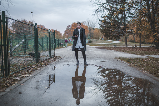 Millennial woman stands in a park next to a pond made of rain and uses a mobile phone, during a cold, autumn day