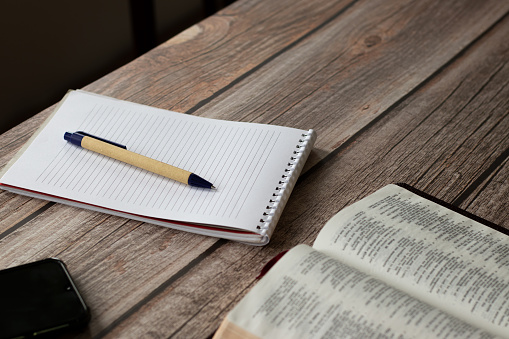 Notebook with a blank page, pen, an open Holy Bible Book, and a mobile phone on a wooden table. Scripture study and writing. Christian biblical concept. A closeup.