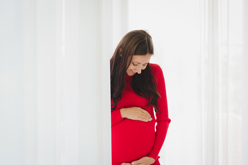 Portrait of pregnant woman in red sweater dress holding her belly with smile and happiness in modern bedroom.