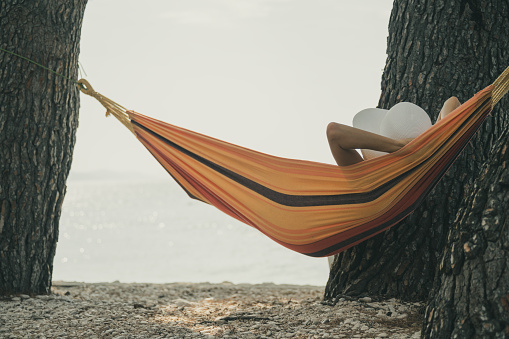 Woman with a summer hat lying in the hammock between the trees on a beach,  relaxing with her arms behind her head