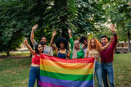 A diverse multiethnic and multicultural group of people are standing outdoors holding a pride flag and waving.