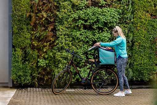 A full length side view of a delivery driver who is checking to make sure that everything in her order is correct. She is loading it into a thermal rucksack resting on her electric bike in Newcastle upon Tyne in the North East of England.