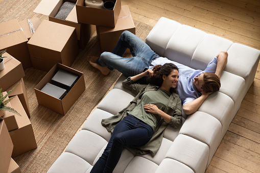 Smiling millennial family couple resting on cozy couch, renewing energy after moving cardboard boxes in new apartment, planning future decorations or discussing interior ideas together at home.