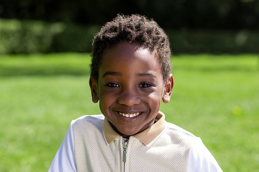 Waist up portrait of a young male boy looking into camera. He is in a public park on a sunny day wearing casual clothing.