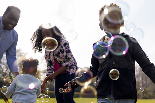Close up of a family of four standing in a public park on a sunny day smiling and laughing catching bubbles. They are all wearing casual clothing.