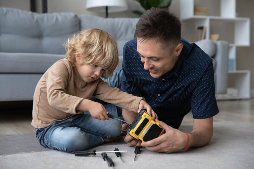 Funny adorable small child boy learning repairing toy car with screwdriver, fixing favorite automobile with caring young father, enjoying spending playtime weekend activity together in living room.