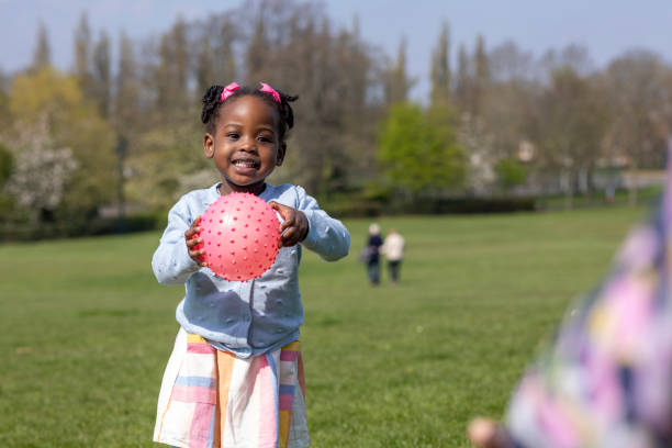 A female toddler holding a bright pink ball in a public park on a sunny day. She is smiling and looking away from the camera wearing casual clothing.