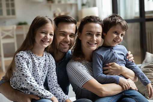 Head shot portrait happy family with two children hugging, sitting on couch in living room at home, smiling young father and mother with little son and daughter enjoying leisure time together