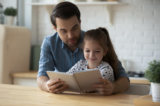 Caring Caucasian father with 9s daughter reading book together, sitting at wooden table in kitchen, loving young dad teaching adorable child girl to read, engaged in educational activity at home