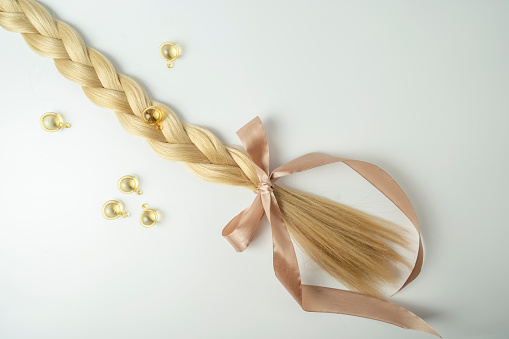 Natural blonde hair and essential oil for hair treatment in golden capsules lying on a white background