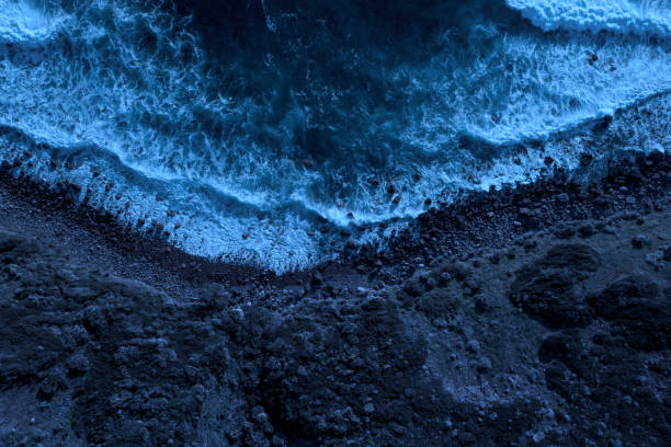 Moonlight at beach: sea waves splashing on waterfront Sea waves seen from above at night, while splashing and flowing on a rocky beach illuminated by moonlight. Dark blue and cold hues. Aerial view. spume stock pictures, royalty-free photos & images