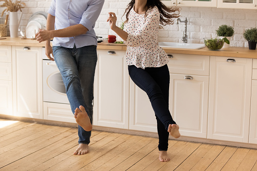 Focus on barefoot legs of happy young family couple dancing to music on warm wooden floor in modern renovated kitchen, joyful millennial man and woman homeowners having fun together on weekend.