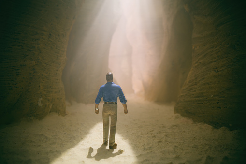 Here we see a silhouetted figure exploring a deserted slot canyon, with a thin beam of sunlight streaming through from overhead. Miniature photography.
