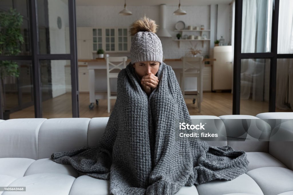 Latina female shiver at freezing flat in warm cap blanket Frozen. Sad latina female sit on couch at freezing cooled studio flat in warm cap and blanket shiver tremble with cold. Unhappy young lady spend time at home feel bad suffer of heating system problems Cold Temperature Stock Photo