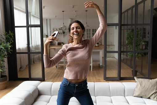 Enjoying freedom. Energetic active hispanic female feel excited crazy jump dance at modern studio apartment listen to music on smartphone. Happy young woman having fun at home celebrate event alone
