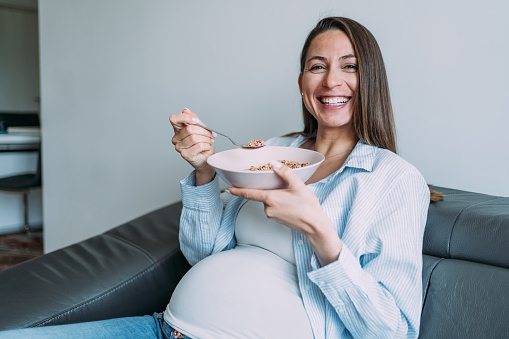 Shot of a beautiful smiling pregnant young woman having healthy breakfast while sitting on the sofa at home.