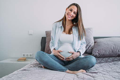 Shot of beautiful pregnant woman relaxing at home while touching her belly. Smiling young expectant mother resting during pregnancy in bedroom.