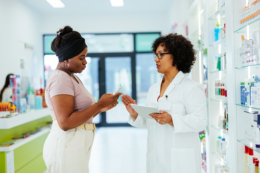 Pharmacy: Professional Black Pharmacist Helping Beautiful Latin Female Customer with Medicine Recommendation, Advice, Talking. Drugstore with Full of Drugs, Pills, Health Care, Beauty Product Packages