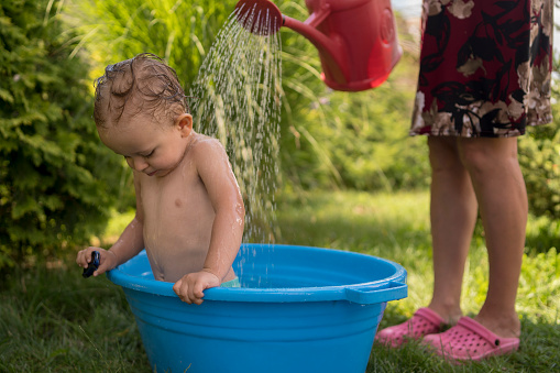 Baby bathes in a small plastic tub in the garden. Kid having fun, splash water and laughs. Summer, vacation, health.