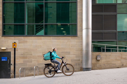 A side view of a female delivery driver cycling alongside an office block looking for the entrance where she is delivering hot food on her electric bike in Newcastle upon Tyne city center.