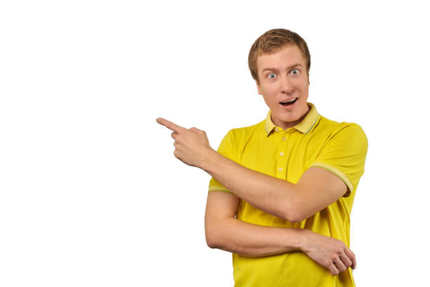 Surprised guy with funny face in yellow T-shirt pointing finger to left, white background stock photo