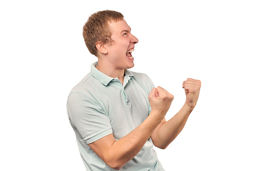 Successful smiling guy in light gray T-shirt rejoices success, isolated on white background, copy space. Funny yelling man celebrating success, excited emotion of triumphant achievement