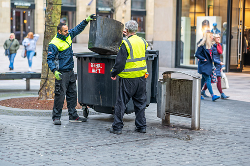 Municipal workers in hi-viz uniforms emptying the trash cans in Cardiff city centre. Pedestrians are pictured walking by