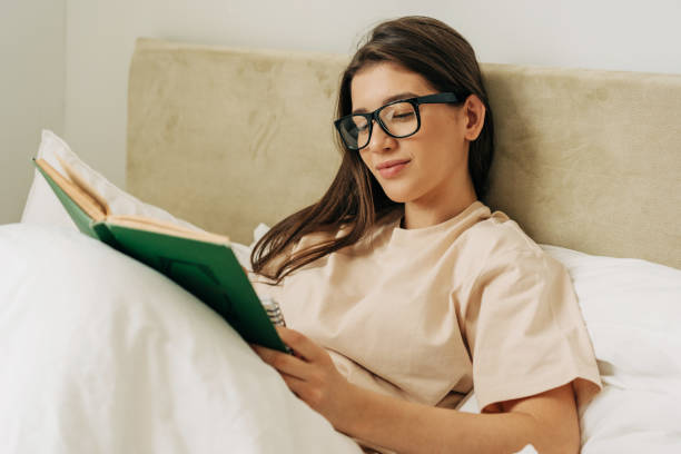 istock Smart student girl in glasses reading a book while lying in bed 1405654408