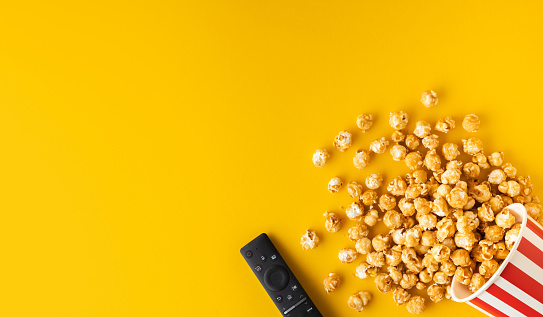 Caramel popcorn scattered from a red and white bucket with remote control on a yellow background. Movie or cinema concept. Flat lay. Top view. Copy space
