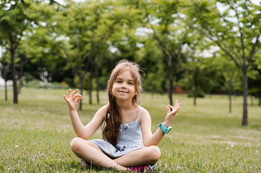 Adorable child girl is sitting and meditate in lotus pose in the park. Relaxation and meditation of kids outdoor. Lifestyle of calm toddler. Enjoy life