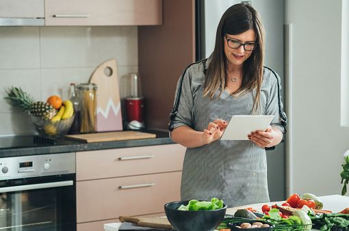 Happy housewife using digital tablet for watching internet tutorials and making healthy meal while standing in front of the table full with fresh organic vegetables at home.