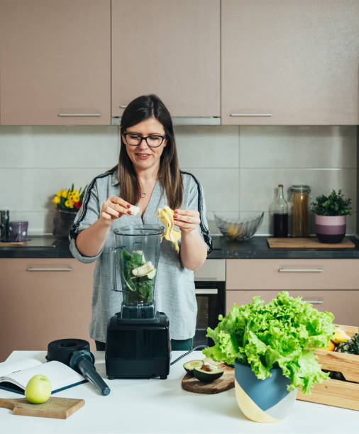 smiling woman making green smoothie for breakfast at home - blender apple banana color image imagens e fotografias de stock