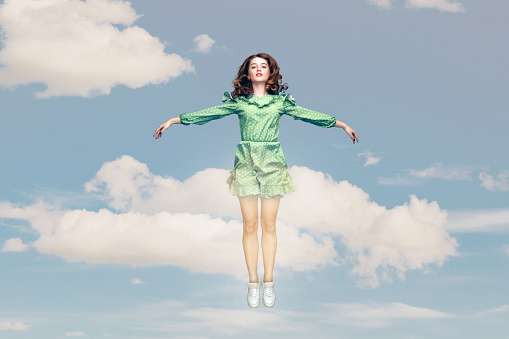 Full length happy calm pretty girl in ruffle dress levitating hovering in mid-air with raised hands as wings, jumping trampoline or flying up in the sky. collage composition on day cloudy blue sky