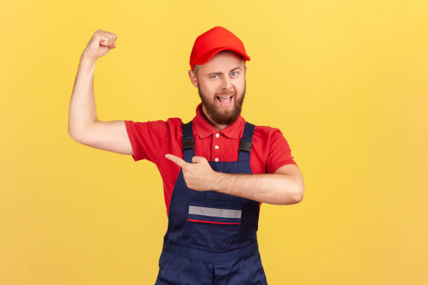 istock Strong worker man wearing blue uniform and red cap standing with raised arms and pointing at biceps. 1405670049