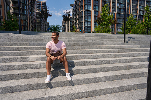 Smiling pleased young man sitting on concrete staircase and looking into distance