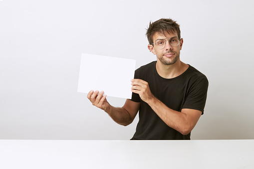 Young Caucasian man with glasses proudly showing his diploma on white background and white desk. He is wearing a black T-shirt.