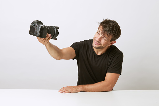 Young Caucasian photographer taking a self photo with a professional camera while smiling. Wear a black t-shirt. Lying on white table, on white background