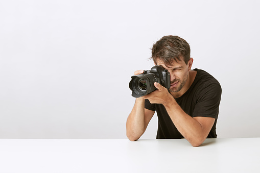Young photographer taking photo with professional reflex camera on white background. Leaning on white desk. Wearing ner t-shirt.