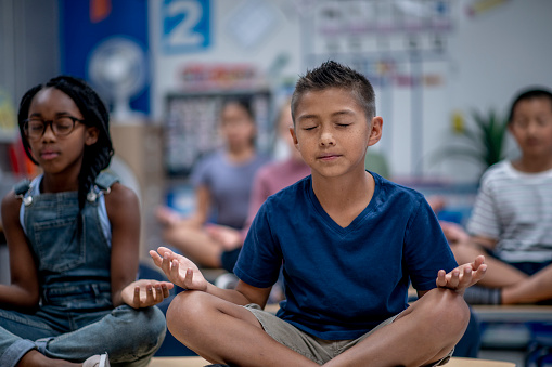 A small group of Elementary students sit cross-legged on the floor with their hands on their knees as they meditate.  They are each dressed casually nd have their eyes closed as they focus on their breathing.