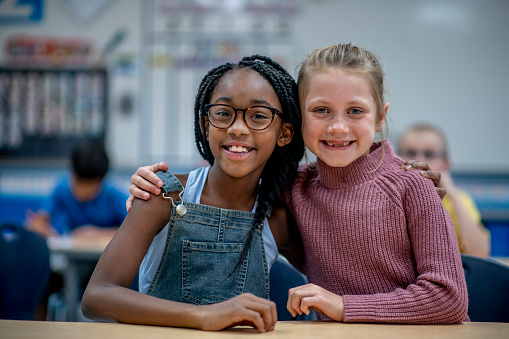 Two female Elementary students sit at a desk, side-by-side and huddled in closely as they pose for a portrait together.  They are both dressed casually and are smiling for the camera.