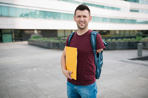 Portrait of a happy young adult male university student with an amputated arm standing in front of the university building, looking at the camera and holding books in his hand