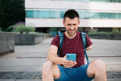 Happy male university student with physical disability sitting on the steps and using a smart phone with his one hand